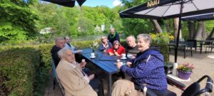 Seniorinnen und Senioren sitzen gemeinsam an einem Tisch im Freien und genießen Kaffee und Eis vor einer idyllischen Teichlandschaft.