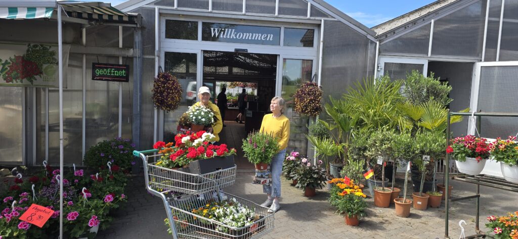 Zwei Kundinnen verlassen eine Gärtnerei mit einem Einkaufswagen voller Blumen und Pflanzen.