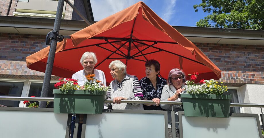 Vier Seniorinnen stehen auf einem Balkon und betrachten gemeinsam bunte Blumen in Balkonkästen.